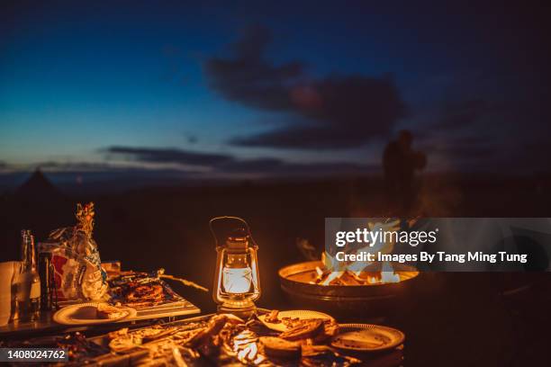 food & oil lamp on a camping table with burning campfire next to it at night in camping fields - campfire no people stock pictures, royalty-free photos & images