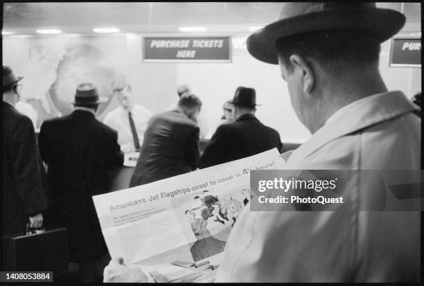View of the shoulder of an unidentified man as he reads a broadsheet advertisement while he stands in line at a ticket counter in National Airport ,...