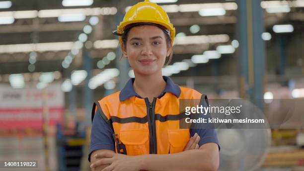 portrait of professional female worker wearing hard hat and safety vest, smiling crossed arm and looking at the camera, standing in the retail warehouse full of shelves with goods cartons on selective rack. employee working on product distribution center - hat rack bildbanksfoton och bilder