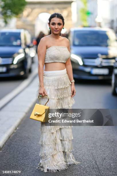 Guest wears green earrings, a beige ribbed / shoulder-off / cropped top, high waist beige fringed wide legs pants, a green and gold ring, a gold...