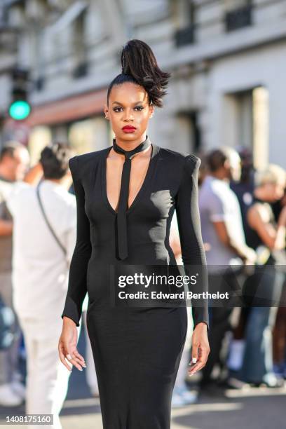 Guest wears diamonds earrings, a black tie, a black V-neck / long sleeves / shoulder-pads / long mermaid dress, outside Jean-Paul Gaultier, during...