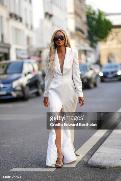 Guest wears black sunglasses, gold earrings, a white V-neck / epaulets / shoulder-pads long slit dress, gold rings, dark brown shiny leather strappy...