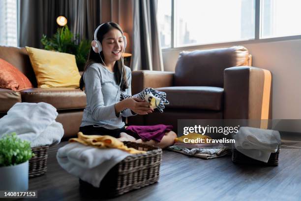 southeast asian woman folding their cloth on weekend while listening music from headphone a young housewife woman touch a s tack of fresh washed clean linen with hands in living room at home - domestic chores stock pictures, royalty-free photos & images