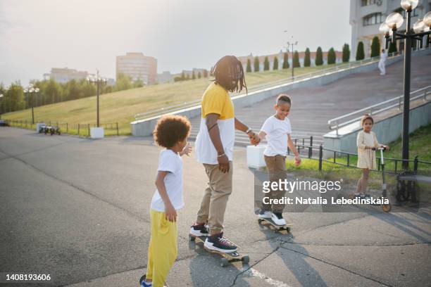 dad with dreadlocks rides skateboards and scooters with children - skaten familie stock-fotos und bilder