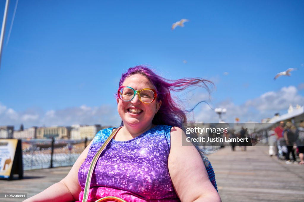 Candid portrait of a young woman who is a wheelchair user smiling while out enjoying a sunny day on a boardwalk by the seaside. She is wearing colorful clothes and her colorful hair is blowing in the breeze.