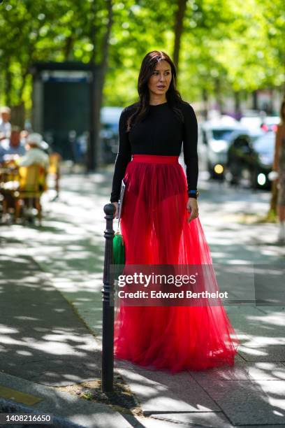 Guest wears a black long sleeves body, a red pleated / tulle long skirt, a green fringed clutch, pumps heels shoes, a blue shiny leather watch,...