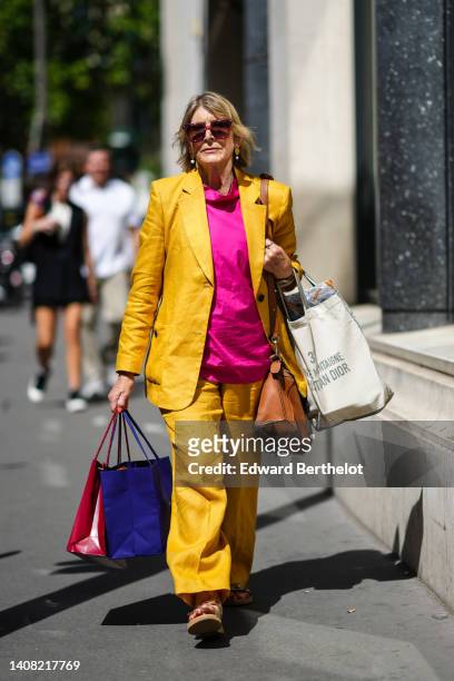 Guest wears pink square sunglasses, gold and pear pendant earrings, gold chain necklaces, a neon pink t-shirt, a yellow mustard blazer jacket,...