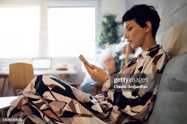 modern young woman using mobile phone while smoking vape - vape cigarette stock pictures, royalty-free photos & images