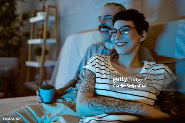 young couple sitting on the sofa and laughing while watching tv - watching television stock pictures, royalty-free photos & images
