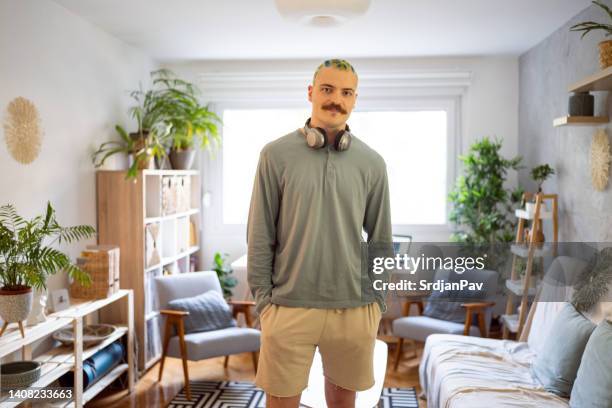 portrait of an urban young man with dyed hair and moustache standing proudly in his room - man living room stockfoto's en -beelden