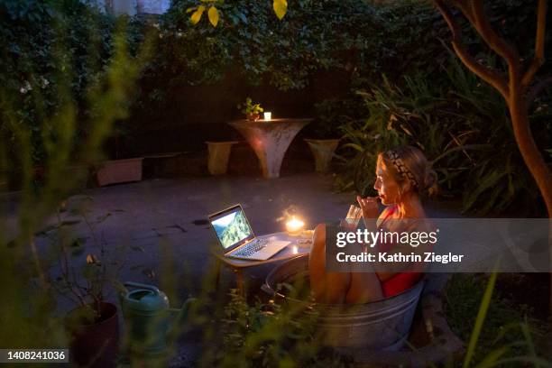 woman cooling off in small tin tub in the garden, watching a movie on laptop - hot summer nights film stock-fotos und bilder