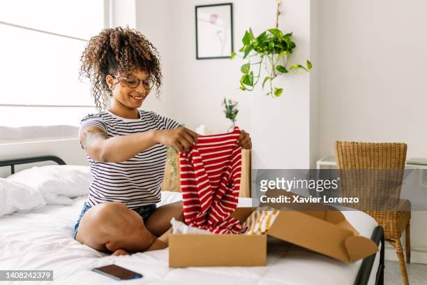 young african woman receiving and opening a parcel box with a clothing item from an online purchase at home. online shopping and shipping service concept. - online shopping opening package stockfoto's en -beelden