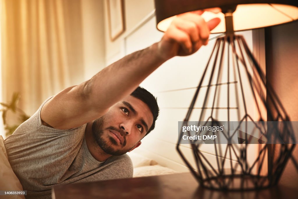 Male feeling exhausted and tired after a long day while turning off a lamp. A young man is switching off the light and going to sleep peacefully under a warm duvet blanket in a cozy bed in a bedroom