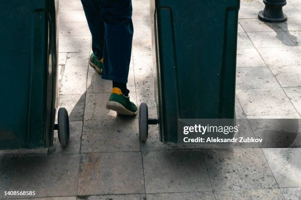 a cleaner or a janitor in dirty shoes collects garbage. a male handyman or scavenger transports black plastic trash cans or dumpsters, tanks on wheels, on the street. the concept of environmental protection, sustainable lifestyle. recycling of garbage. - müllmann stock-fotos und bilder