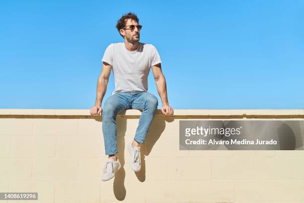 young man dressed in casual clothes sitting on yellow brick wall with blue sky in background. - european outdoor urban walls stockfoto's en -beelden