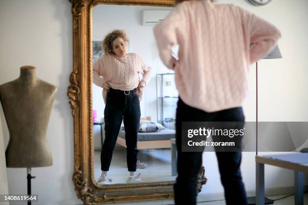 young woman with curly hair standing in front of full length mirror at home, pulling jeans up - woman full length mirror stock pictures, royalty-free photos & images