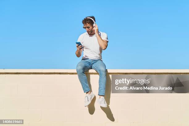 young man dressed in casual clothes using his cell phone while listening to music with wireless headphones, sitting on a yellow colored brick wall with blue sky in the background. - brick phone stock pictures, royalty-free photos & images