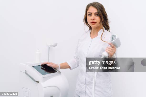 female medical therapist look at camera and hold handpiece head of laser hair removal machine next to her on white background - electrolysis stock pictures, royalty-free photos & images