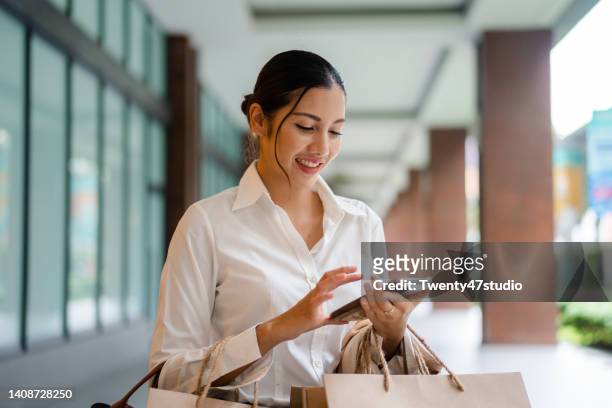 asian woman with shopping bags using a smartphone while walking in the city - shopping center stock-fotos und bilder
