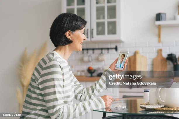 smiling senior woman having online consultation with female physician on smartphone in kitchen. telemedicine concept. telecommuting from home during pandemic. seeking for help and complaining about symptoms, horizontal - ehealth stockfoto's en -beelden