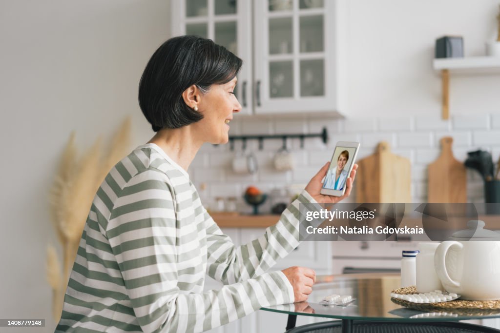 Smiling senior woman having online consultation with female physician on smartphone in kitchen. Telemedicine concept. Telecommuting from home during pandemic. Seeking for help and complaining about symptoms, horizontal