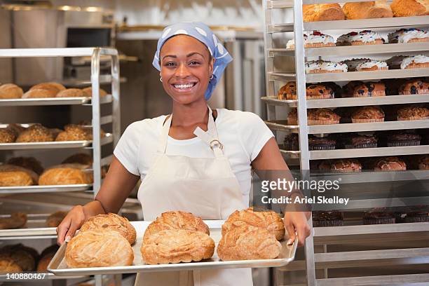 african american baker holding tray of bread in bakery - baker man stock-fotos und bilder