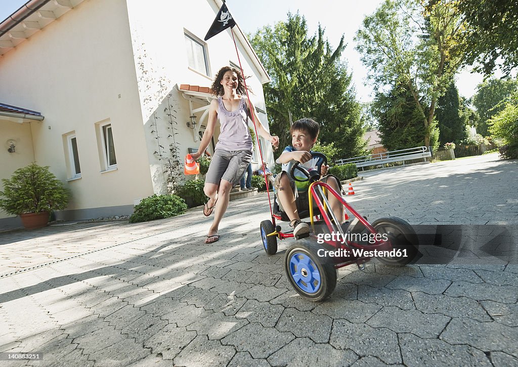 Germany, Bavaria, Boy driving pedal go kart and woman running with family in background