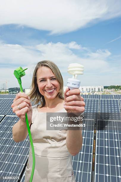 germany, munich, woman holding electric cord and lightbulb in solar energy, smiling, portrait - energy efficient lightbulb stock-fotos und bilder