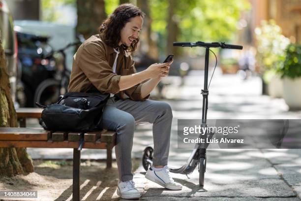 young man sitting on bench and using mobile phone in the city - mobilität stock-fotos und bilder