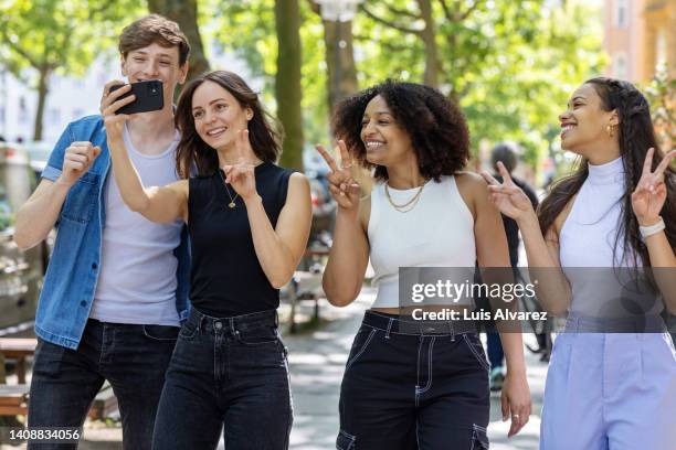 cheerful woman taking selfie with multi-ethnic friends in city - victory sign stock-fotos und bilder