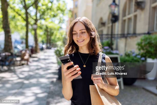 smiling young woman with smartphone walking on the street - sehen stock-fotos und bilder