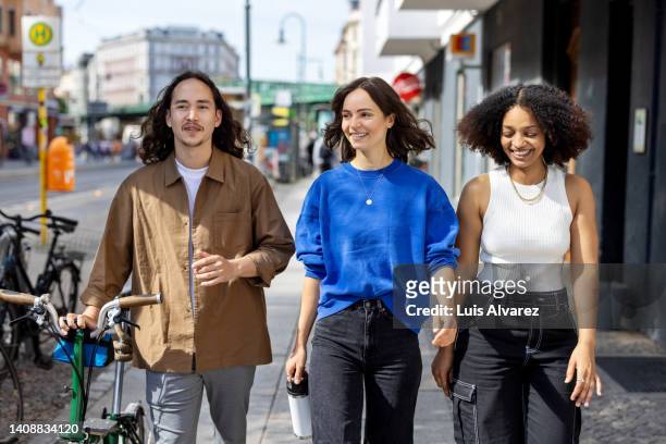 happy young people walking down the city street with a bicycle and smiling - drei frauen stock-fotos und bilder