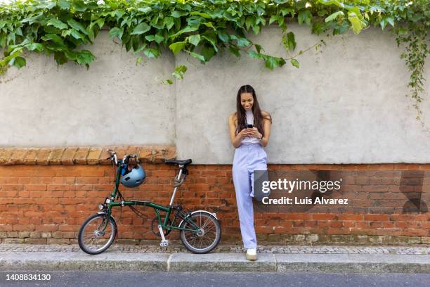 young woman leaning to a wall and texting on her phone with her bicycle on side - purple pants - fotografias e filmes do acervo