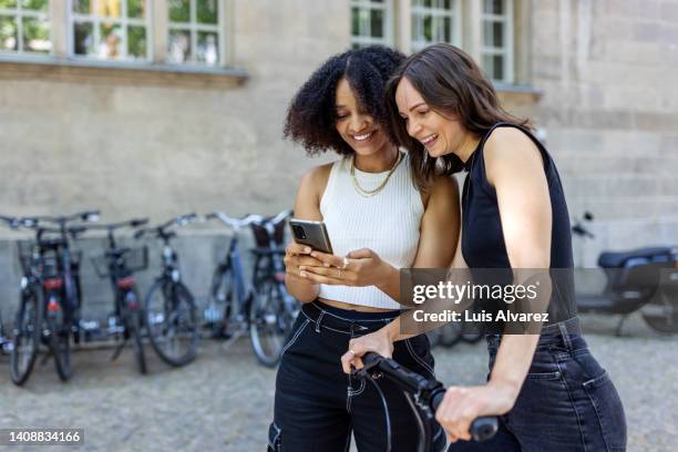 woman showing her phone to a female friend with bicycle on city street - smartphone friends stock-fotos und bilder