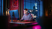 Young Handsome Man in Headphones Using Computer in Stylish Loft Apartment in the Evening. Creative Male Smiling, Working from Home, Browsing Videos on Social Media. Urban City View from Big Window.