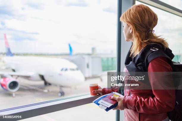 mulher parada na sala de espera do portão com documentos de viagem e café para ir, olhando pela janela - passagem de avião - fotografias e filmes do acervo