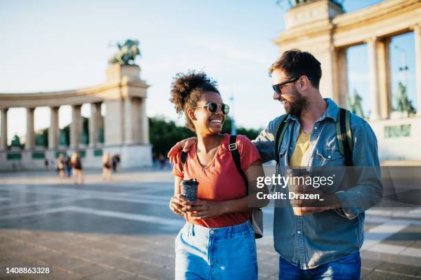 beautiful young multiracial couple enjoying city break, holding coffee cups and wearing backpacks - boedapest stockfoto's en -beelden