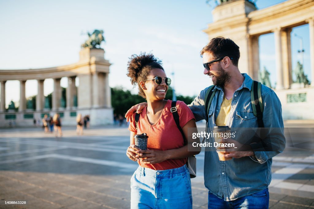 Beautiful young multiracial couple enjoying city break, holding coffee cups and wearing backpacks