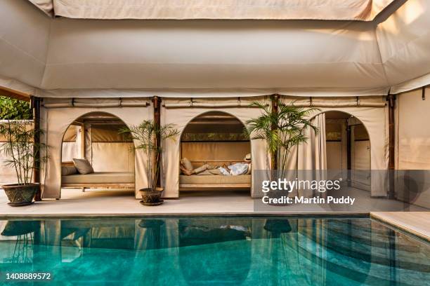 mature man relaxing on couch  next to salt water pool with arched tent, at eco friendly luxury glamping resort in bali. - luxury tent stock pictures, royalty-free photos & images