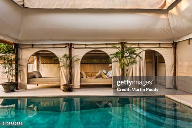 mature man reading while relaxing on couch, next to salt water pool with arched tent, at eco friendly luxury glamping resort in bali. - luxury travel stock pictures, royalty-free photos & images