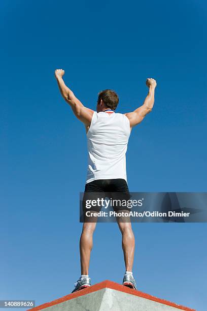 male athlete standing on winner's podium with arms raised in victory, rear view - olympic podium stock pictures, royalty-free photos & images