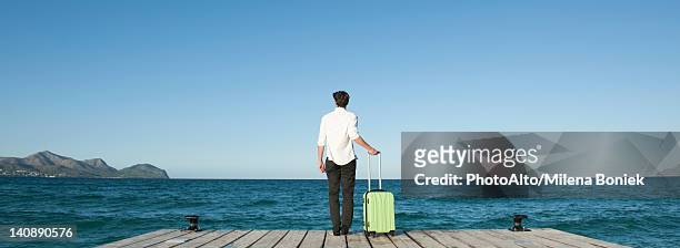 man standing on dock with suitcase, looking at ocean view, rear view - pier 1 stock pictures, royalty-free photos & images