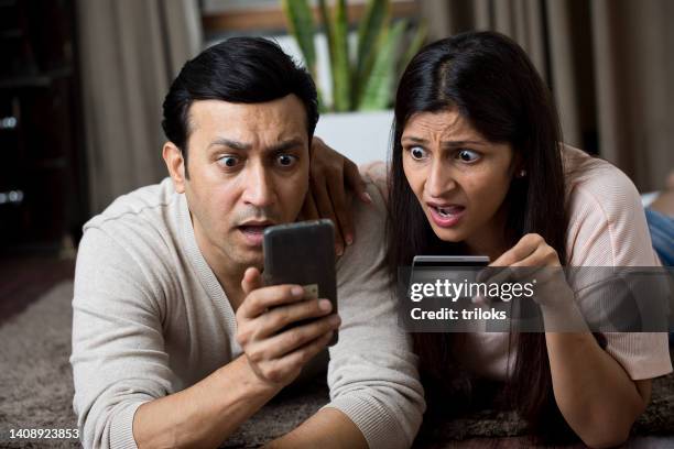 shocked couple looking worried while using mobile phone and credit card at home - fraudulent stock pictures, royalty-free photos & images