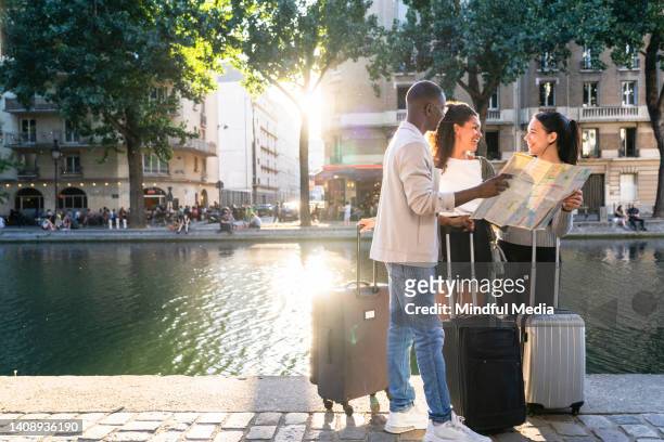 full body shot of group of young people looking at map and standing next to a river with suitcases - information kiosk stock pictures, royalty-free photos & images