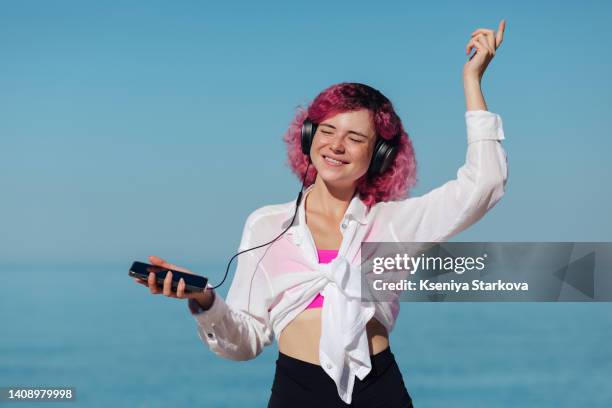 a young woman with freckles and short colored pink hair in a white shirt and a pink top against a blue sky on the beach listens to music from a phone with headphones - wavy hair beach stock pictures, royalty-free photos & images