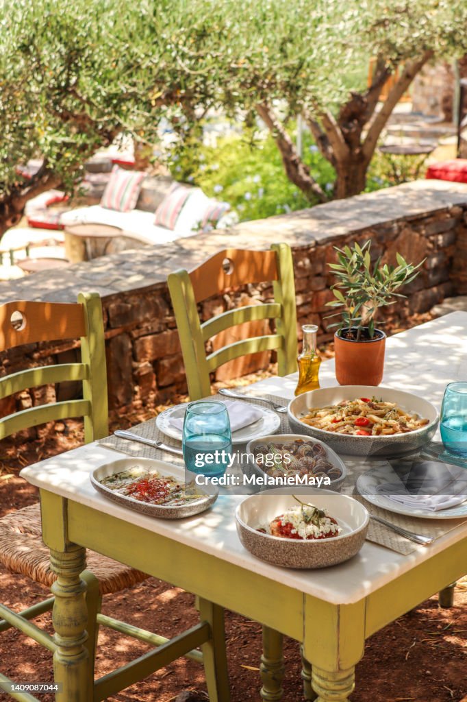 Griechischer Esstisch mit Essen und Tellern unter Olivenbäumen mit kretischen Köstlichkeiten