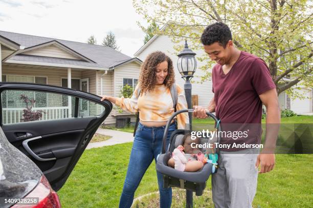 young parents putting baby daughter into car outside their home - baby car seat stock pictures, royalty-free photos & images