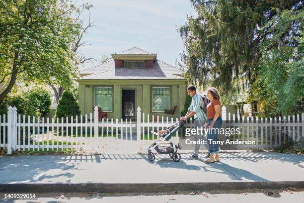 young mother and father walking with baby daughter in stroller on neighborhood sidewalk - district stockfoto's en -beelden