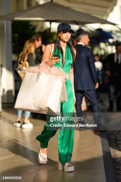 Guest wears a black cap, a green satin shiny V-neck / glowing jumpsuit, a beige large shopping bag, white latte leather strappy / block heels sandals...