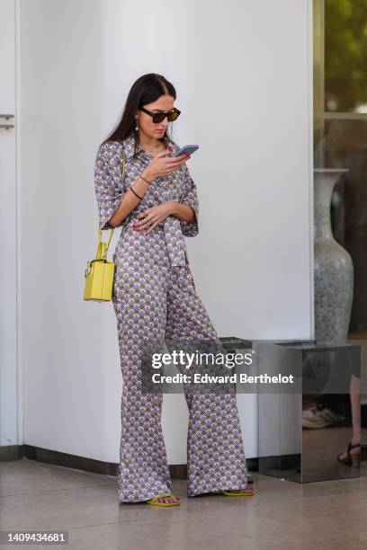 Guest wears black sunglasses, gold and pendant pearl earrings, a white latte with purple and green print pattern belted jumpsuit, a yellow shiny...